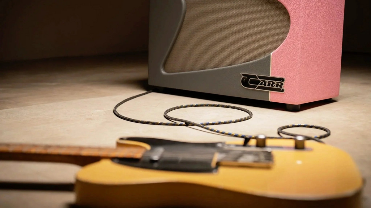 A Carr guitar amplifier in pink, gray, and black, shown with a yellow electric guitar in the foreground on a wooden floor.