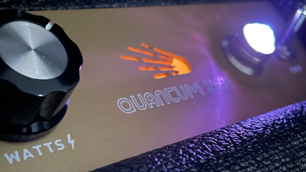 Close-up of a guitar amplifier control panel with a black rotary knob labeled WATTS, a gold faceplate, an orange hand logo, and the text QUANTUM ZEN, with a bright illuminated light on the right.